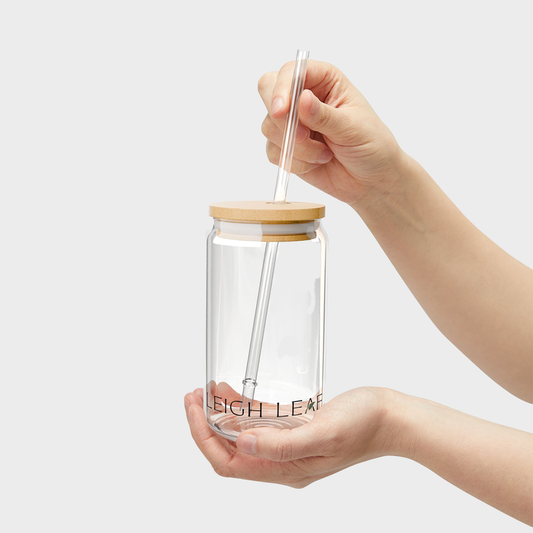 Clear glass tumbler with wooden lid and straw held by a hand on a light gray background